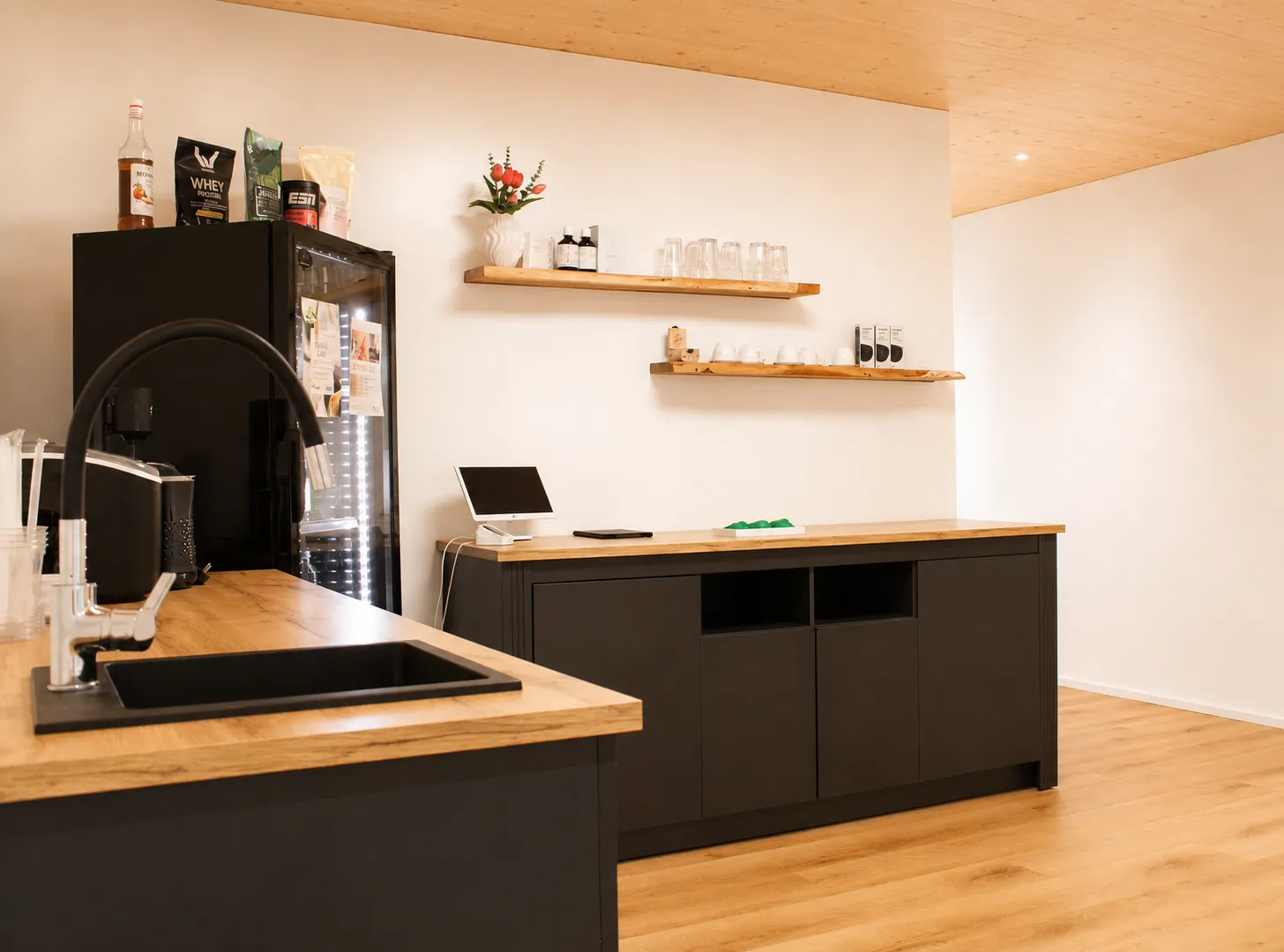 Der helle Thekenbereich mit schwarzen Einbauten, Holzarbeitsplatten, Kühlschrank und offenen Holzregalen.